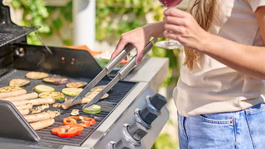 Woman grilling turning sausages and vegetables over with metal tongs. She is holding a glass of red wine, there is green foliage behind her, and she is wearing a loose t-shirt and denim jeans. New pet-friendly apartment homes for rent with private fenced backyards in Arizona, Texas, Florida, and North Carolina with pool, pickleball, dog park, gym, clubhouse and gated community. *No breed or weight restrictions. Yardly