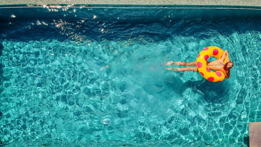 Aerial photo of woman in pool lying on her stomach in a pink polka dot and yellow ring float. The pool water is icy blue and clear. New pet-friendly apartment homes for rent with private fenced backyards in Arizona, Texas, Florida, and North Carolina with pool, pickleball, dog park, gym, clubhouse and gated community. *No breed or weight restrictions. Yardly