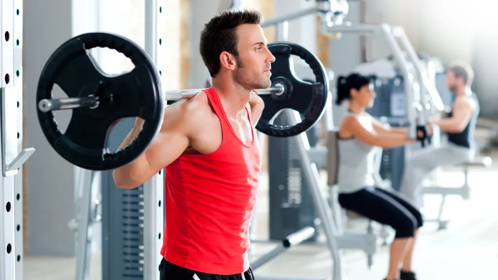 Man in weightroom wearing a red shirt and black shorts with squat bar across his back preparing to squat. In the background you can see a man and a woman on other weight machines and a new clean gym. New pet-friendly apartment homes for rent with private fenced backyards in Arizona, Texas, Florida, and North Carolina with pool, pickleball, dog park, gym, clubhouse and gated community. *No breed or weight restrictions. Yardly