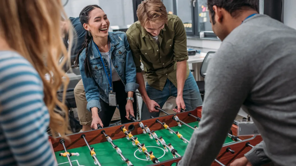 Men and women playing foosball in the game area of a clubhouse. One woman is laughing and the man beside her is looking down intrigued and focused on the game while another man and woman are in the foreground also participating from the other side of the table. New pet-friendly apartment homes for rent with private fenced backyards in Arizona, Texas, Florida, and North Carolina with pool, pickleball, dog park, gym, clubhouse and gated community. *No breed or weight restrictions. Yardly