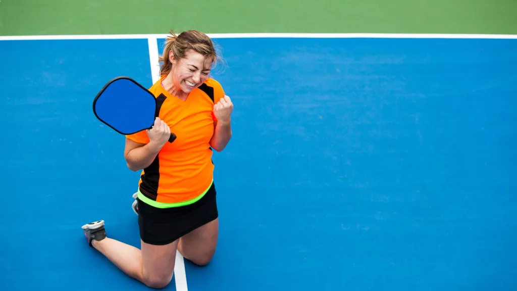 Girl wincing in victorious pose on her knees on a pickleball court after winning pickleball tournament. she has her racket in one hand and her other is a fist held closely to her. She is wearing an orange and black athletic tee and tennis skirt and sneakers. New pet-friendly apartment homes for rent with private fenced backyards in Arizona, Texas, Florida, and North Carolina with pool, pickleball, dog park, gym, clubhouse and gated community. *No breed or weight restrictions. Yardly